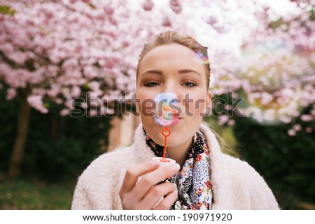 Portrait of beautiful young woman blowing bubbles at park. Pretty female model with bubble wand at spring blossom garden.