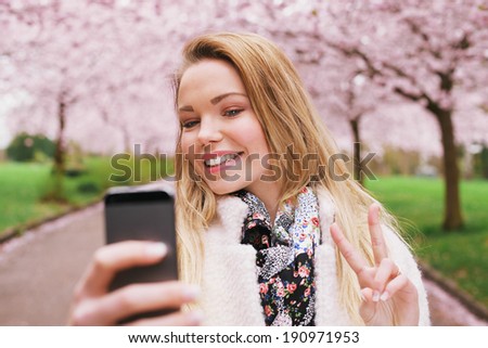 Pretty young female model gesturing peace sign while taking her picture with cell phone. Beautiful caucasian young woman taking self portrait while at spring blossom park.