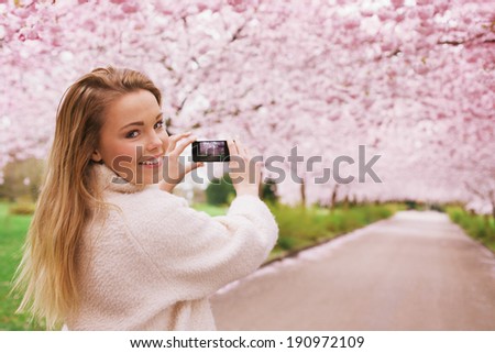 Young woman using her smartphone to capture images of the path and cherry blossoms tree at park, Young female looking over her shoulder while taking pictures with her phone at spring blossom garden.