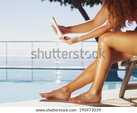 Woman sitting on deck chair by the swimming pool applying sun cream onto her legs Female model sunbathing at the poolside Woman sitting on deck chair by the swimming pool applying sun cream onto her legs Female model sunbathing at the poolside