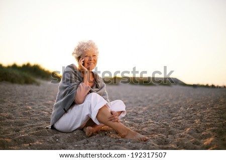 Relaxed retired woman wearing shawl sitting on sandy beach making a phone call. Old caucasian woman sitting on the beach looking at camera outdoors