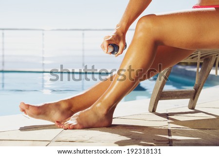 Woman applying suntan spray onto her legs. Female sitting on recliner chair by the swimming pool sunbathing.