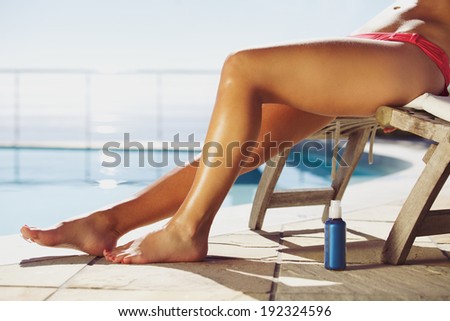 Young female relaxing on lounge chair with a suntan spray by the pool. Young lady sunbathing by the swimming pool.