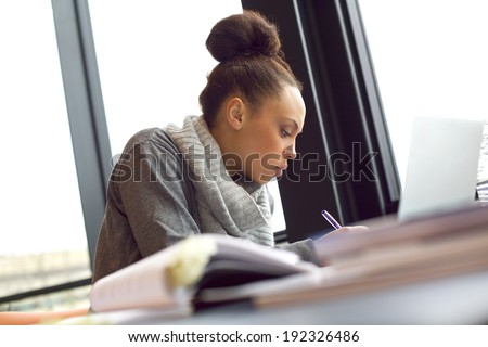 Young african american woman taking notes from books for her study. Sitting at table with books and laptop for finding information.