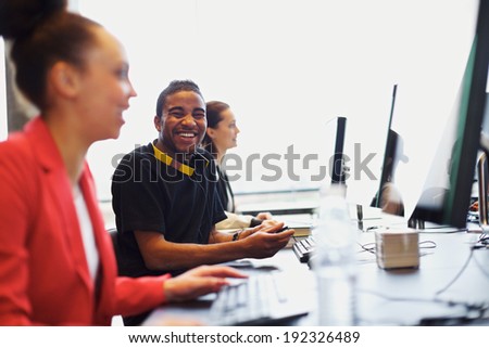 Young afro american man looking at camera smiling while working on computer in modern classroom. Young students sitting at college computer lab.