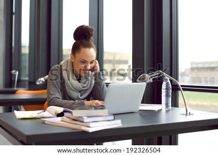 Pretty young woman sitting in library using laptop smiling. Young woman sitting at table surfing internet for notes for her study.