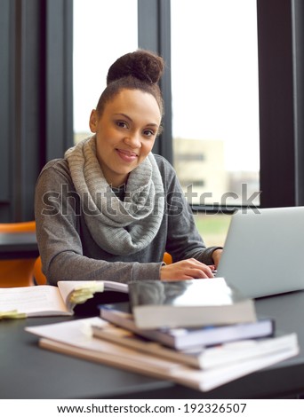 Happy young african american woman student with laptop and books sitting at table looking at camera smiling. Female student studying in library.