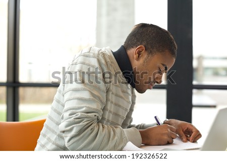 Young afro american man taking notes from books for his study. Student sitting at table with books for finding information.