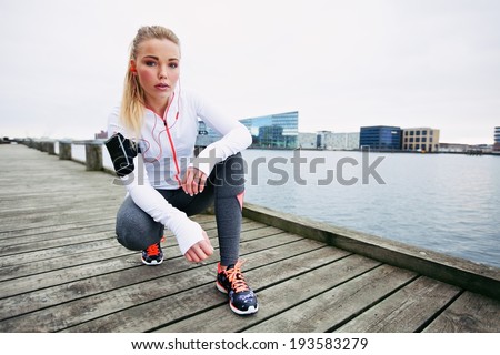 Young woman runner resting after running workout. Female fitness model crouching on sidewalk along river. Female jogger taking a break.