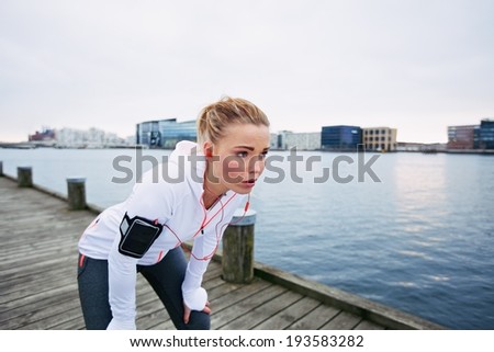 Female runner standing bent over and catching her breath after a running session along river. Young woman taking break after a run.