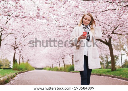 Young lady at spring park listening music from her mobile phone. Relaxed woman taking a walk at spring blossom garden listening music.