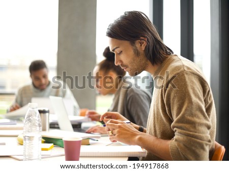 Young man talking notes for study with students studying in background. University students preparing for final exams in library.