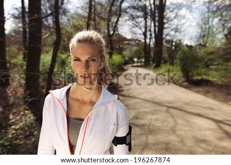 Portrait of pretty young woman in sports clothing wearing earphones while on fitness routine in forest. Young female runner of forest trail.