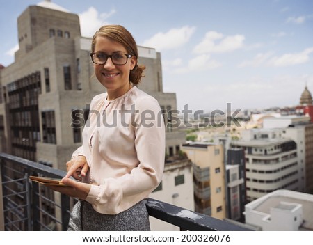 Pretty young woman standing in balcony holding digital tablet looking at camera smiling. Caucasian female in terrace with view of city in background.