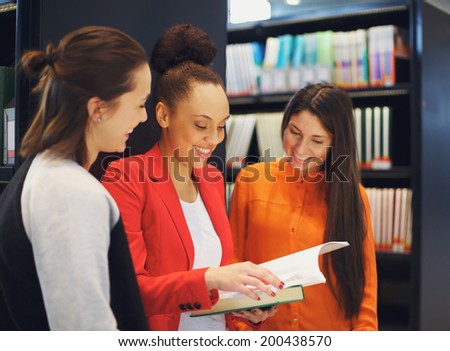 Diverse group of college students in library reading a book together. Female students looking for information in reference books for their studies.