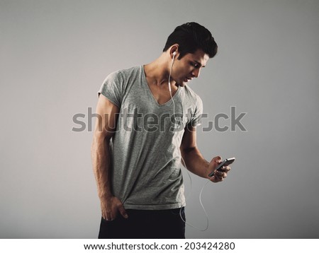 Handsome young man in casual t-shirt listening music on cell phone. Hispanic male model wearing earphones and listening to music through his smart phone on grey background.