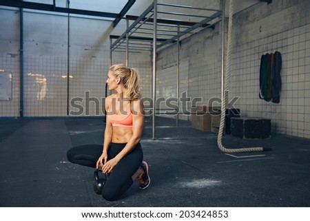 Young woman kneeling on floor with kettle bell and looking away. Fit caucasian female athlete at crossfit gym relaxing after tough workout.