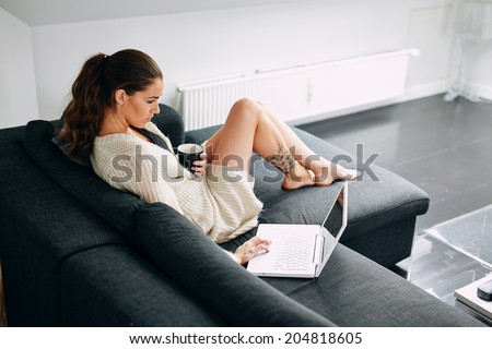 Portrait of young woman sitting on sofa checking her emails. Attractive female model with a cup of coffee and laptop at home in morning.
