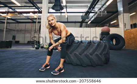 Beautiful and strong young woman sitting on a big tire after her exercise. Woman taking break after crossfit workout at gym.
