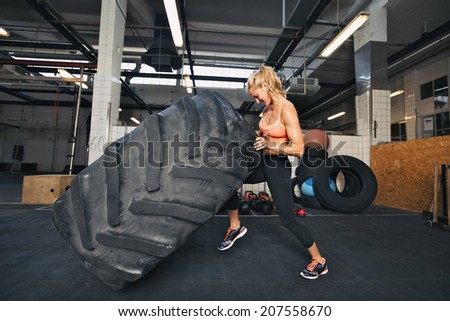 Muscular young woman flipping tire at gym. Fit female athlete performing a tire flip at crossfit gym.