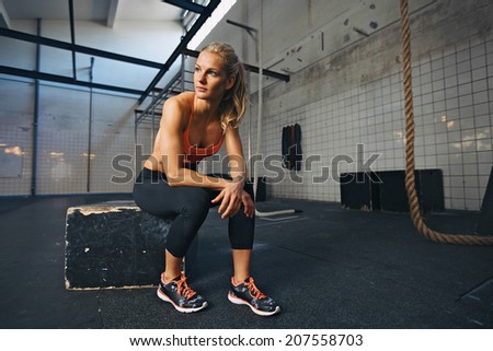 Young woman sitting on a box at gym after her workout. Caucasian female athlete taking rest after exercising at gym.