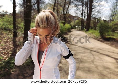 Image of pretty young blonde wearing earphones to listen music before starting her running workout. Fit female athlete before her run in forest.