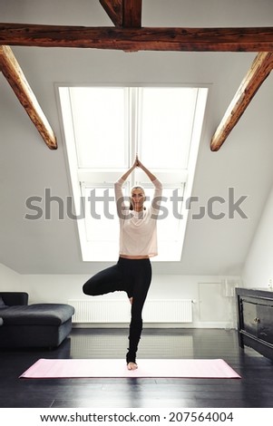 Young woman practicing yoga in living room. Healthy female standing on one leg and meditation at home. Fit female in tree pose doing yoga indoors.