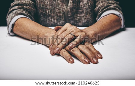 Close-up studio shot of a senior woman\'s hands resting on grey surface. Old lady sitting with her hands clasped on a table.