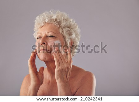 Portrait of senior woman applying lotion on her face against grey background. Mature caucasian woman applying anti-wrinkle face cream.