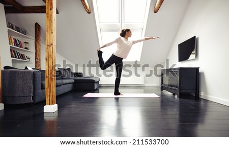 Full length image of fit young lady practicing yoga at home. Healthy young woman exercising in living room. Caucasian female model in warrior pose.