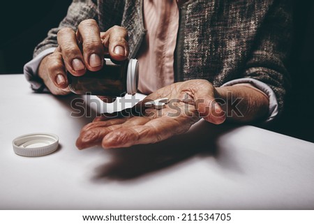 Cropped shot of a old woman sitting at a table shaking a pill out of a pill bottle. Focus on hands. Senior female taking medicine.