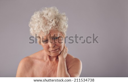 Studio shot of senior woman suffering from neck pain against grey background. Caucasian old woman in discomfort with sore neck.