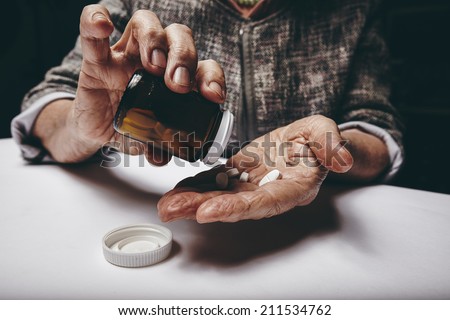 Cropped view of elderly woman taking prescription medicine from pill bottle. Senior female\'s hands pouring pills on her palm while sitting at a table.