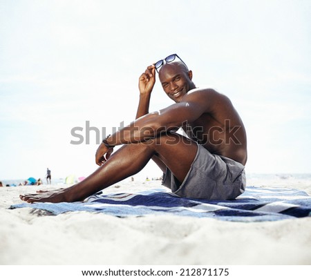 Portrait of handsome young man sitting on beach looking at camera smiling. Shirtless muscular man sunbathing on beach.