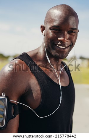 Image of happy young man wearing earphones looking at camera smiling. Fit young african male model outdoors. Muscular male runner outdoors.