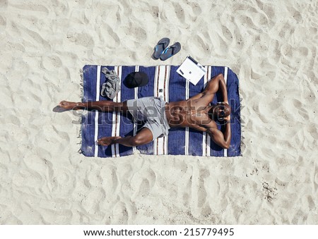 Top view of afro american young man relaxing on beach. Muscular guy wearing sunglasses and listening to music on headphones lying on a beach mat. Shirtless male model sunbathing.