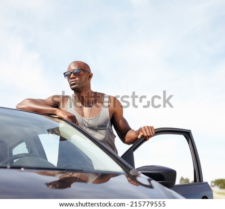 Portrait of smart young man leaning on car. African male model wearing sunglasses standing by his car looking away with door open outdoors.