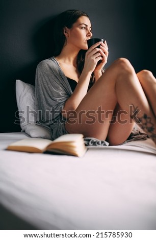 Young lady sitting on bed holding a cup of coffee looking away thinking. Female model in night wear drinking coffee.
