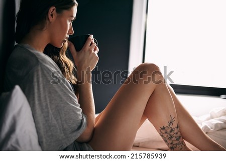 Young woman looking away while drinking a cup of coffee in bedroom. Caucasian female model drinking coffee while sitting on bed.