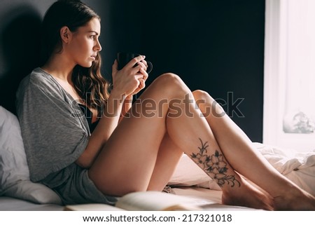 Young woman holding a cup of coffee and looking away thoughtfully while on bed. Attractive female model drinking coffee in bedroom.