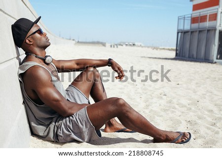 Man sitting on beach leaning to a sea wall. African male model wearing sunglasses, cap and headphones relaxing on beach.
