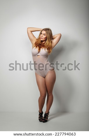 Full length studio image of beautiful voluptuous woman in body stockings standing on grey background. Sexy young plus size female model posing in underwear.