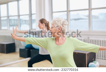 Two women doing stretching and yoga workout at gym. Female trainer in background with senior woman in front during physical training session