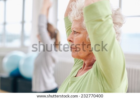 Close-up image of senior woman practicing yoga at gym. Active senior woman exercising at health club with female trainer in background.