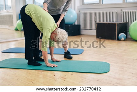 Senior woman bending forward and touching her toes being helped by gym instructor. Elder woman doing back exercise with help from physical therapist at gym.