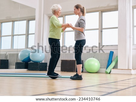Female trainer helping senior woman standing on a balance board at gym. Elder woman exercising being assisted by personal trainer.