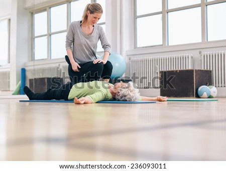 Female physical therapist assisting senior woman with leg exercise in therapy gym. Elder woman lying on floor being assisted by personal trainer in stretching exercises.