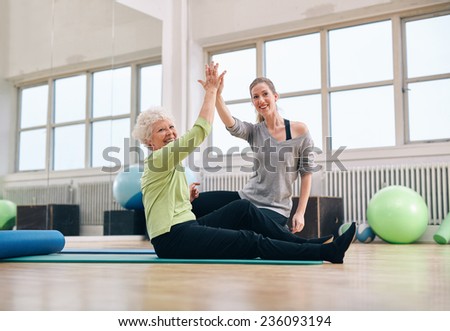 Senior woman sitting on fitness mat and her personal trainer joining hands at gym. Fitness women giving high-five at health club.