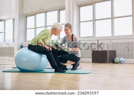 Elderly woman in a gym sitting on exercise ball and talking to her personal female trainer about exercise plan. Senior woman and coach looking at health report together.