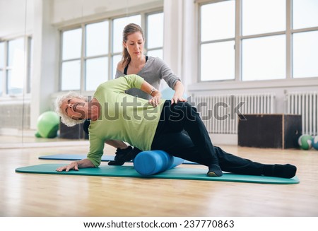 Female instructor helping senior woman using a foam roller for a myofascial release massage at gym.